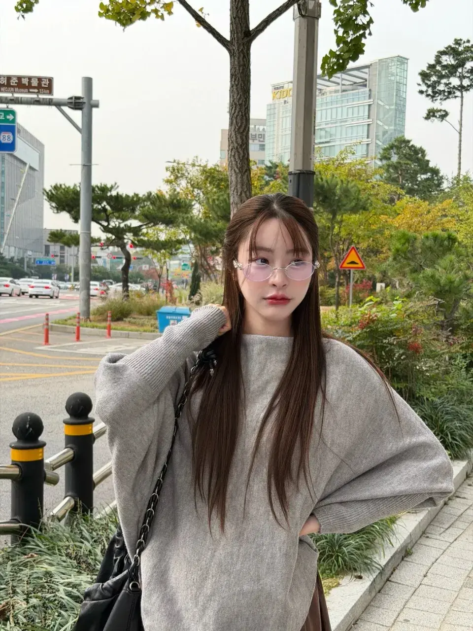 ใส่แว่นกรอบใส เสื้อสเวตเตอร์สีเทาตัวโคร่ง ยืนริมถนนที่ป้ายเป็นภาษาเกาหลี ใบไม้เริ่มเปลี่ยนสีแล้ว