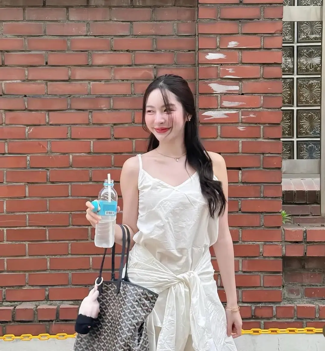 ชุดเดรสสีขาว มือหนึ่งถือขวดน้ำ อีกมือหิ้วกระเป๋าใบใหญ่ ยืนหน้ากำแพงอิฐแดง