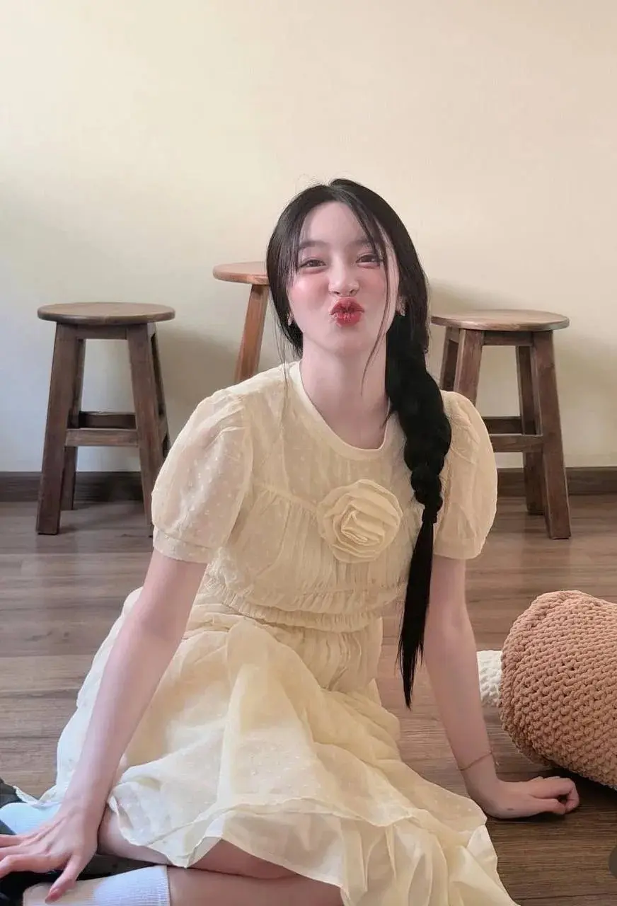 นั่งพื้นไม้ในชุดเดรสสีครีม มีดอกไม้ผ้าติดอก ข้างหลังมีเก้าอี้ไม้สองตัว