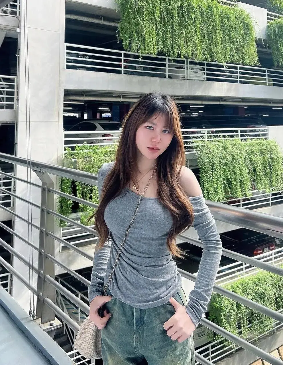 เสื้อแขนยาวสีเทาเปิดไหล่ ยีนส์ทรงหลวม สะพายกระเป๋าสานใบเล็ก ยืนพิงราวระเบียงตึกที่มีต้นไม้ห้อยลงมาเต็มผนัง