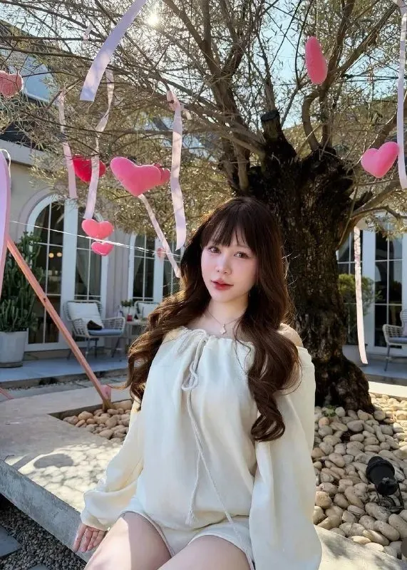 เสื้อเบลาส์สีครีมแขนพอง นั่งใต้ต้นไม้ที่แขวนหัวใจสีชมพูไว้ทั้งต้น